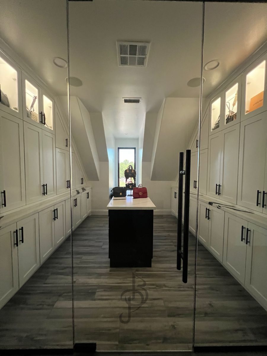 Glass-enclosed walk-in closet with white shaker wardrobes, lit handbag display cabinets, and a black center island